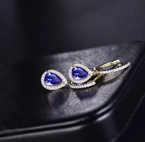 A pair of gold and natural diamond white stone earrings with pear-shaped blue tanzanite gemstones, displayed on a black surface.