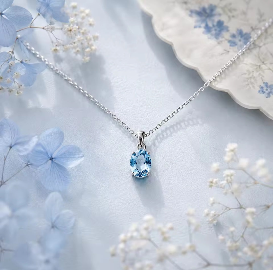 A silver chain necklace with a teardrop-shaped light blue natural blue topaz gemstone pendant, styled on a light blue textured surface with blue hydrangeas and white baby's breath flowers.