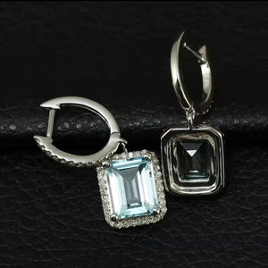 A pair of white gold earrings with rectangular blue topaz emerald-cut light blue gemstones and small clear natural diamonds accent stones, displayed on a textured black surface.