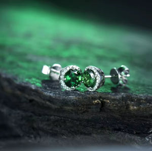 A pair of white gold stud earrings with round green tsavorite gemstones, photographed close-up on a dark textured surface with a blurred green background.