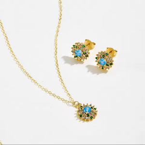 Gold jewellery set featuring a delicate chain necklace and matching stud earrings, each adorned with a central light blue stone surrounded by small clear stones and green accents, displayed on a clean white background.