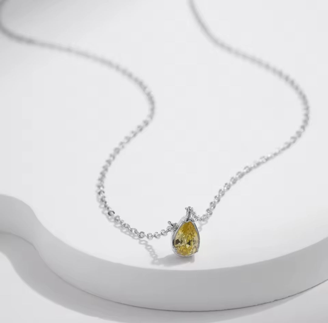 A delicate silver chain necklace featuring a teardrop-shaped yellow gemstone pendant in a silver setting, displayed on a white curved platform.