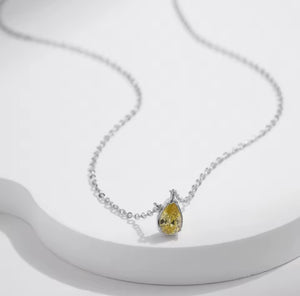 A delicate silver chain necklace featuring a teardrop-shaped yellow gemstone pendant in a silver setting, displayed on a white curved platform.