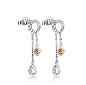 A pair of white gold drop earrings with a circular stud, a short chain, a small square amber/ yellow sapphire gemstone, and a teardrop-shaped silver dangling charm, shown on a white background.