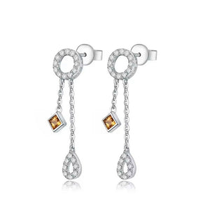 A pair of white gold dangle earrings featuring circular bezel-set clear stones, a small square amber gemstone, and a teardrop-shaped dangling design with clear stones, shown on a white background.