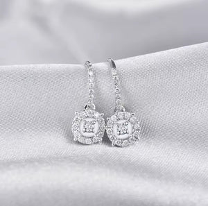 A pair of diamond drop earrings with square halo designs set with clear stones, resting on a textured white fabric background.