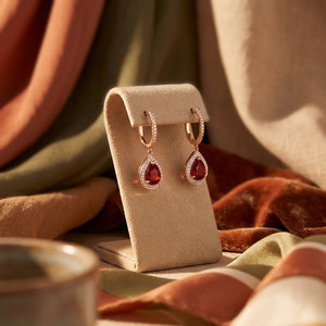 hoop drop earrings with teardrop-shaped red ruby gemstones and small clear  natural diamond stones, displayed on a beige jewellery stand against draped fabric.