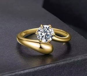 A gold ring with a round-cut clear gemstone in a prong setting, displayed on a dark textured surface.