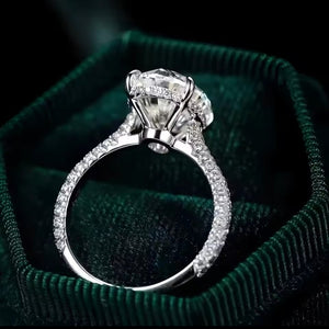 A silver-toned diamond ring with a large round center stone and pavé-set side stones, displayed inside a dark green velvet ring box.
