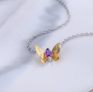 A delicate silver chain necklace featuring a butterfly pendant with textured gold wings and a central purple amethyst gemstone, resting on a white marble surface.