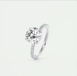 A silver solitaire engagement ring with a round brilliant-cut center stone and pavé-set accent stones along the band, shown on a light gray background.
