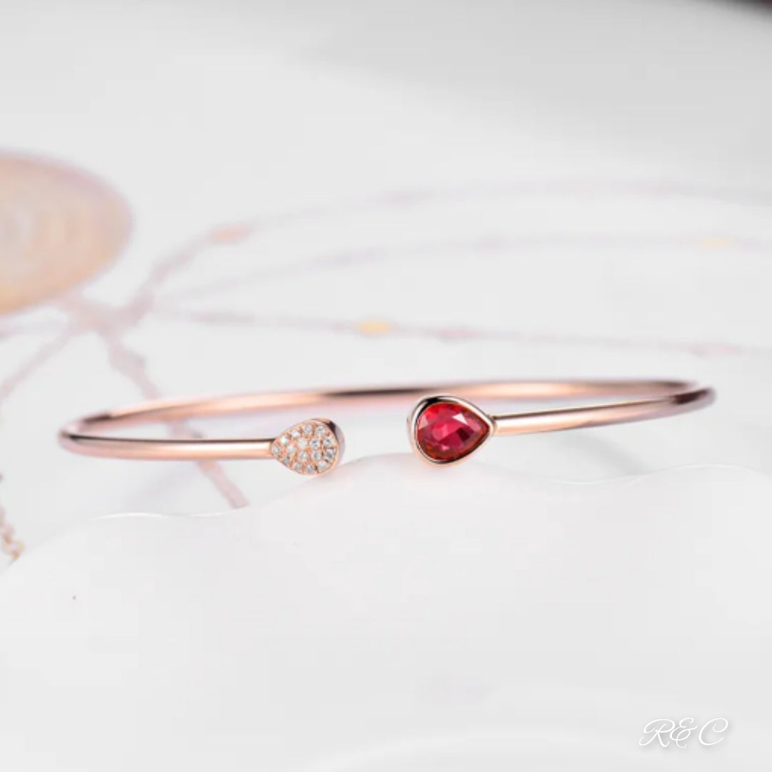 A rose gold bangle bracelet with a central ruby and surrounding diamonds, displayed on a white surface.