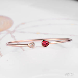 A rose gold bangle bracelet with a central ruby and surrounding diamonds, displayed on a white surface.